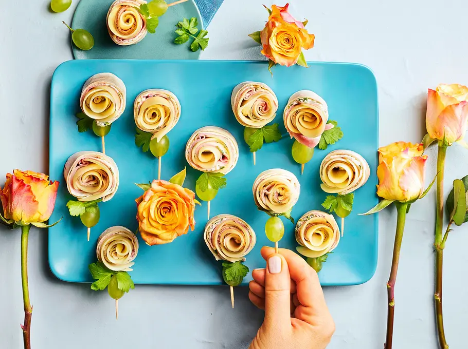 Mehrere Wrap-Röllchen in Rosenform auf einem türkisfarbenen Teller, dekoriert mit grünen Trauben und Petersilie, dazu gelbe Rosen als Dekoration.