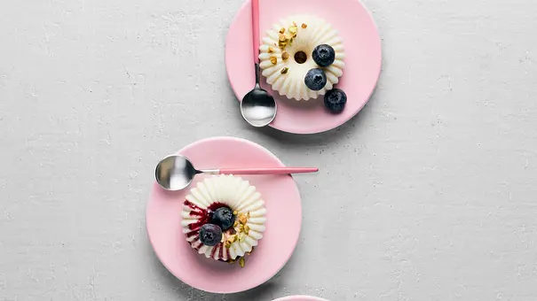 Drei kleine Joghurt-Pannacotta auf rosa Tellern, mit Beerensoße und frischen Blaubeeren dekoriert, daneben auf einem rosa Unterteller eine Schale mit weiteren Blaubeeren. - Foto:  House of Food / StockFood Studios / Meike Bergmann 