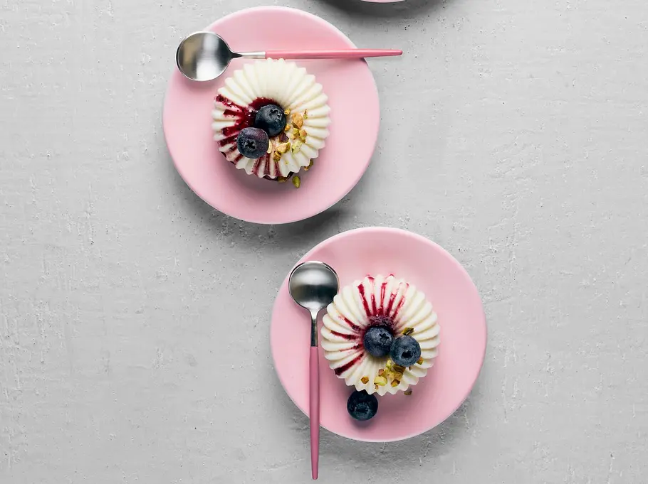 Drei kleine Joghurt-Pannacotta auf rosa Tellern, mit Beerensoße und frischen Blaubeeren dekoriert, daneben auf einem rosa Unterteller eine Schale mit weiteren Blaubeeren.