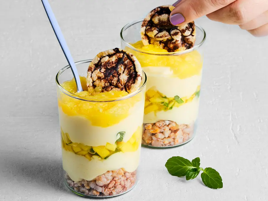 Zwei Gläser mit Mango-Reiswaffel-Trifle, geschichtet mit knusprigen Reiswaffelstücken, Mango und Quarkcreme, getoppt mit einer Schokoladenreiswaffel, serviert auf einer hellen Tischplatte.