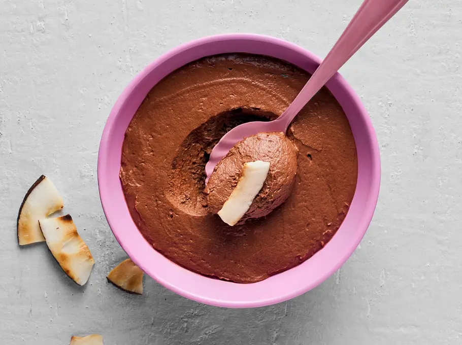 Schoko-Kokos-Mousse Schoko-Kokos-Mousse in einer rosa Schüssel, mit einem Löffel und Kokosstücken garniert, dazu ein paar frische Kokosstücke daneben, im Hintergrund zwei Tassen Cappuccino, eine auf rosa Untertasse.