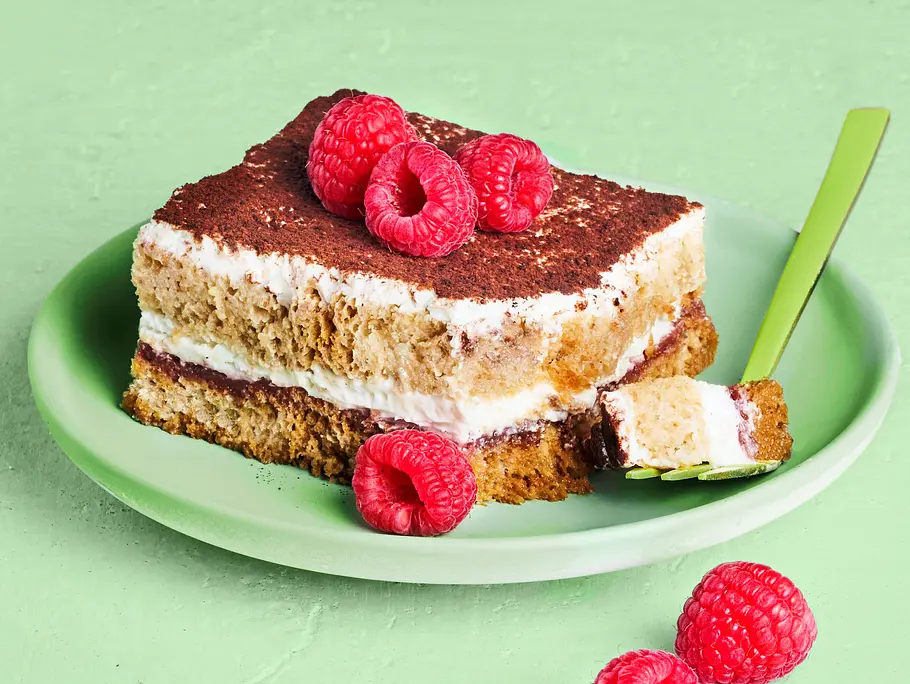 Stück Himbeer-Tiramisu mit Kakaopulver bestäubt und frischen Himbeeren garniert, serviert auf einem grünen Teller mit kleiner Gabel
