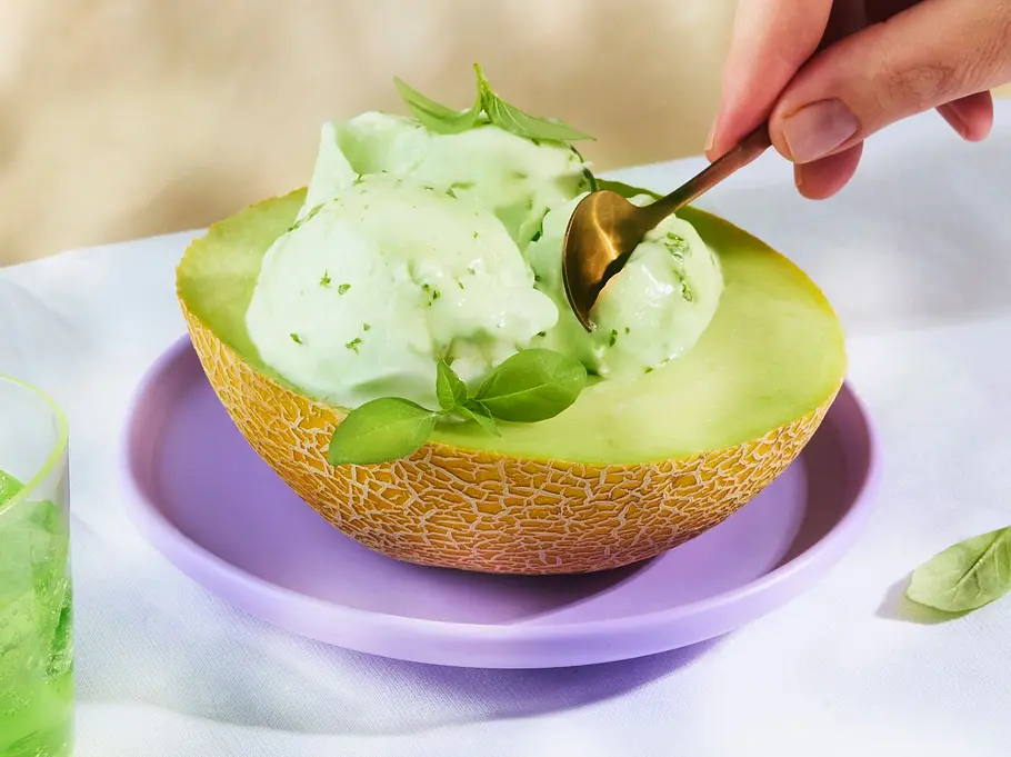 Melonen-Limetten-Sorbet mit Basilikum Drei Kugeln hellgrünes Melonen-Limetten-Sorbet in einer ausgehöhlten Melonenhälfte, angerichtet auf einem fliederfarbenen Teller. Daneben ein Glas grünes Getränk mit Limettenscheibe und Basilikum.