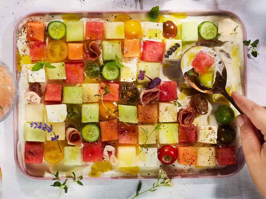 Snackplatte im Schachbrettmuster mit Würfeln aus Melone und Feta, dazu Gurken-, Tomatenstücke und Schinkenröllchen auf cremigem Labneh, serviert auf einem rechteckigen Teller auf weißer Tischdecke. Daneben Gläser und eine Flasche Rosé.