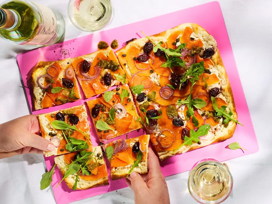 Rechteckiger Flammkuchen mit Melone, Ziegenkäse, roten Zwiebeln, Oliven und Rucola, serviert auf einem pinken Schneidebrett auf weißer Tischdecke. Zwei Gläser Weißwein und eine Flasche Wein daneben.