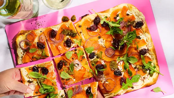 Rechteckiger Flammkuchen mit Melone, Ziegenkäse, roten Zwiebeln, Oliven und Rucola, serviert auf einem pinken Schneidebrett auf weißer Tischdecke. Zwei Gläser Weißwein und ein Weinglas daneben. - Foto: LECKER @ Bauer Media Group / StockFoodStudios/ Meike Bergmann Rechteckiger Flammkuchen mit Melone, Ziegenkäse, roten Zwiebeln, Oliven und Rucola, serviert auf einem pinken Schneidebrett auf weißer Tischdecke. Zwei Gläser Weißwein und ein Weinglas daneben. - Foto: LECKER @ Bauer Media Group / StockFoodStudios/ Meike Bergmann
