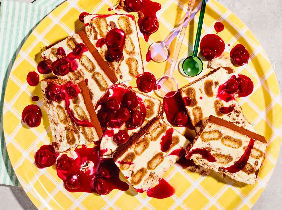 Mehrere Stücke Tiramisu-Semifreddo mit sichtbaren Biskuitschichten und intensiv rotem Kirschkompott auf einer gelb-karierten Platte, ein Stück auf einem lila Teller mit goldfarbenem Löffel, dazu drei Espressotassen.