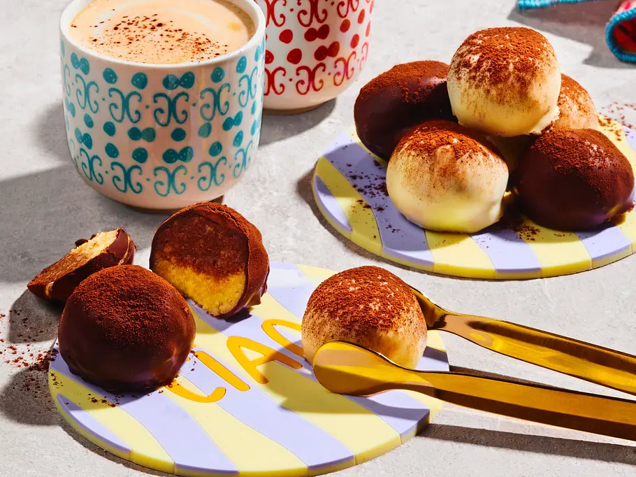 Mehrere schwarz-weiße Tiramisu-Trüffel auf kleinen, gelb-lila gestreiften Tellern, zum Teil mit Kakao bestäubt, dazu zwei gemusterte Tassen Cappuccino und eine goldene Zange auf heller Unterlage.