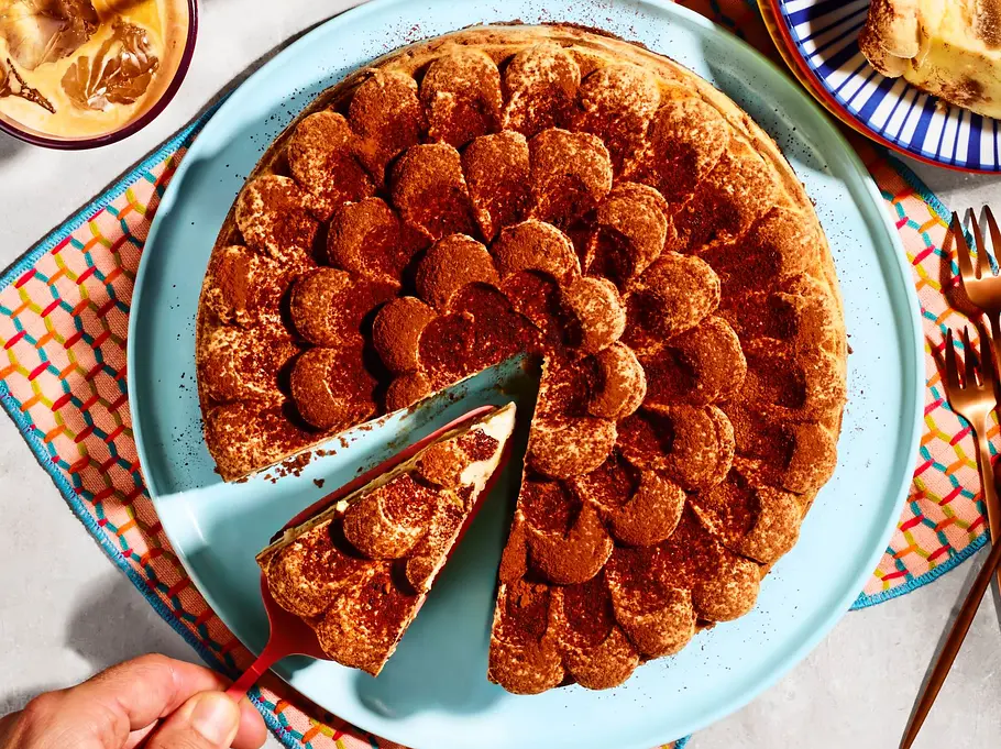 Tiramisu-Cheesecake auf einem hellblauen Teller, mit einer dekorativen Schicht Kakaopulver bestäubt, daneben ein Stück auf einem blauen, gestreiften Teller und zwei Gläser Eiskaffee auf einem bunten Untersetzer.