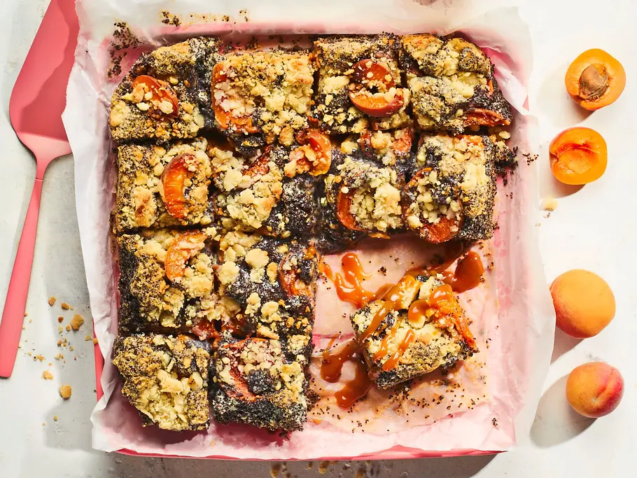 Aprikosen-Mohn-Schnitten mit Marzipanstreuseln Rezept Rechteckige Aprikosen-Mohn-Schnitten mit goldenen Marzipanstreuseln in einer mit rosa Backpapier ausgelegten Form, garniert mit frischen Aprikosen und einem pinkfarbenen Tortenheber.