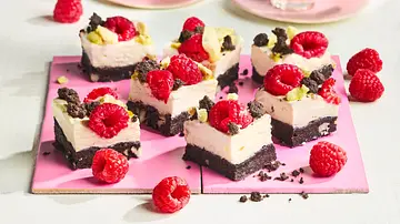 Kleine Cheesecake-Würfel mit cremiger Füllung auf dunklem Schoko-Nuss-Boden, belegt mit frischen Himbeeren und Schokostreuseln, angerichtet auf einer rosa Platte. - Foto: StockFood Studios / Meike Bergmann