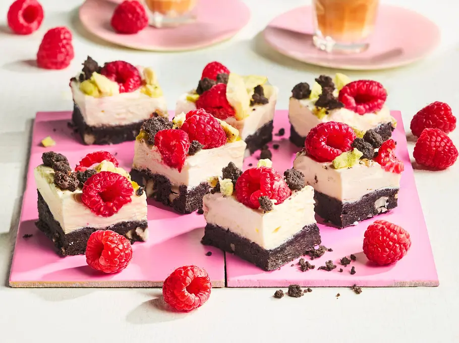 Cheesecake-Würfel mit Schoko-Nuss-Boden Kleine Cheesecake-Würfel mit cremiger Füllung auf dunklem Schoko-Nuss-Boden, belegt mit frischen Himbeeren und Schokostreuseln, angerichtet auf einer rosa Platte.