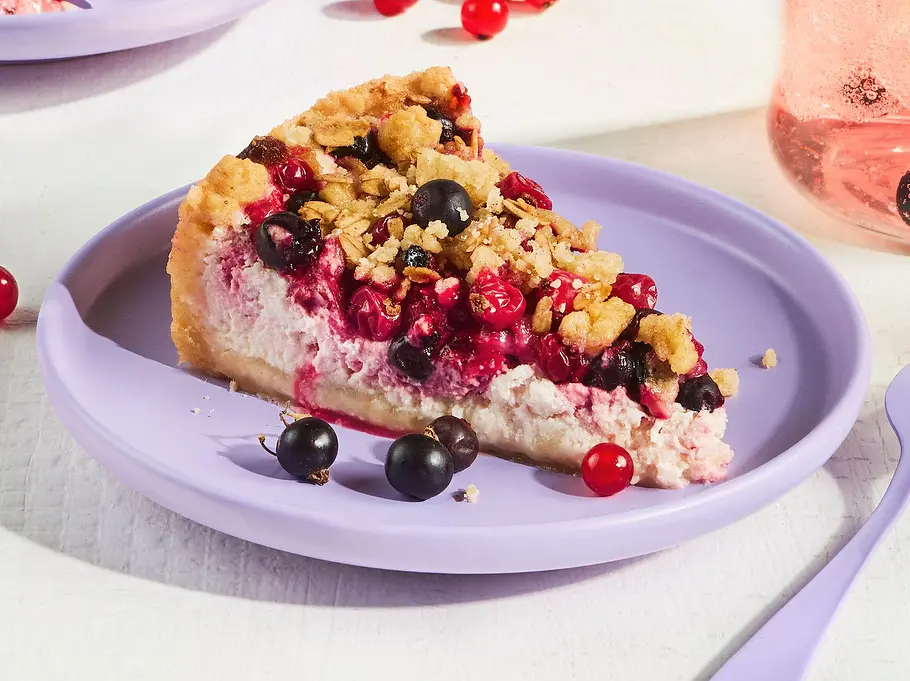 Johannisbeerkuchen mit Schmand und Lillet-Kick Rezept Drei Stücke Johannisbeerkuchen mit Schmandcreme und goldenen Streuseln auf fliederfarbenen Tellern, dekoriert mit frischen roten und schwarzen Johannisbeeren, daneben ein Glas Lillet-Drink.