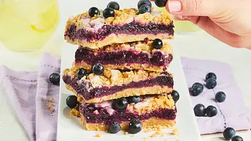 Vier Heidelbeer-Prasselkuchen-Stücke mit knusprigen Streuseln, gefüllt mit dunkler Beerenmasse, auf einem weißen rechteckigen Teller, garniert mit frischen Heidelbeeren, daneben ein hellviolettes Stofftuch. - Foto: StockFood Studios / Meike Bergmann