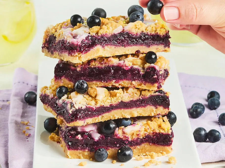 Heidelbeer-Prasselkuchen mit Limoncello Rezept Vier Heidelbeer-Prasselkuchen-Stücke mit knusprigen Streuseln, gefüllt mit dunkler Beerenmasse, auf einem weißen rechteckigen Teller, garniert mit frischen Heidelbeeren, daneben ein hellviolettes Stofftuch.