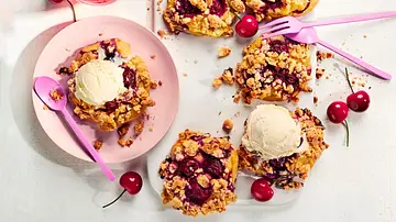 Mehrere Streuseltaler mit saftigen Kirschen und goldbraunen Streuseln auf einer weißen Platte, einige mit einer Kugel Grießpudding dekoriert, daneben frische Kirschen und rosafarbene Teller und Besteck. - Foto: StockFood Studios / Meike Bergmann Mehrere Streuseltaler mit saftigen Kirschen und goldbraunen Streuseln auf einer weißen Platte, einige mit einer Kugel Grießpudding dekoriert, daneben frische Kirschen und rosafarbene Teller und Besteck. - Foto: StockFood Studios / Meike Bergmann