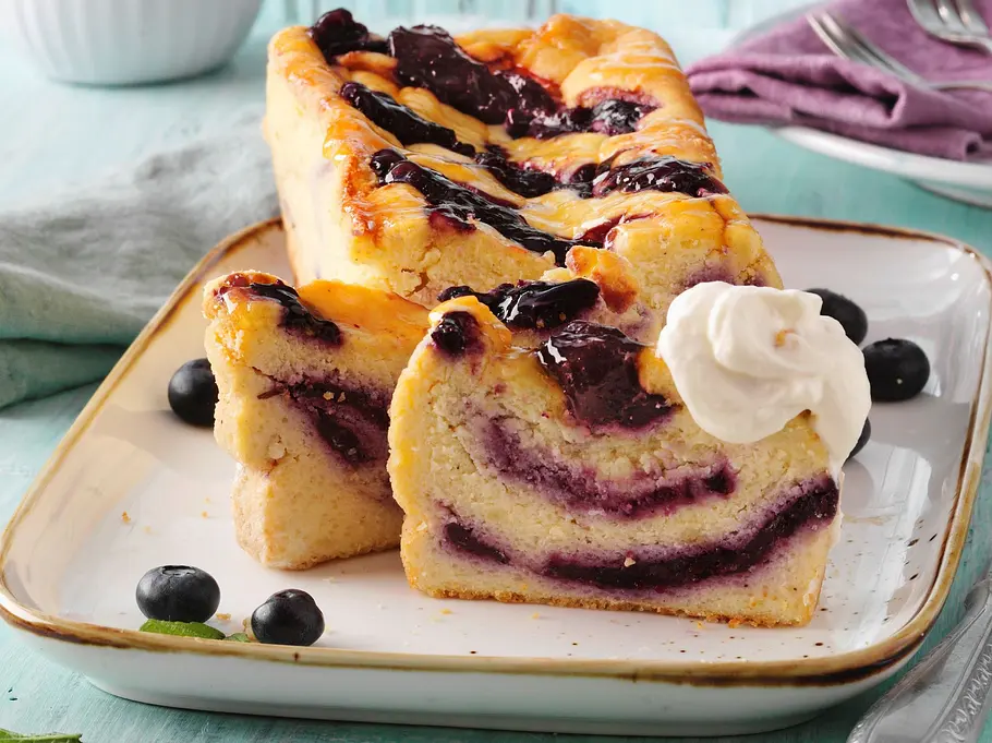 Ein Heidelbeer‑Käsekuchen wird in Scheiben geschnitten und mit frischen Beeren sowie einem Klecks Sahne serviert.