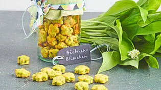 Kleine, gebackene Bärlauchkekse in Blümchenform, dekorativ in ein Glas mit grünem Stoffdeckel gefüllt, daneben frischer Bärlauch auf einer dunklen Oberfläche. - Foto: LECKER @ Bauer Media Group