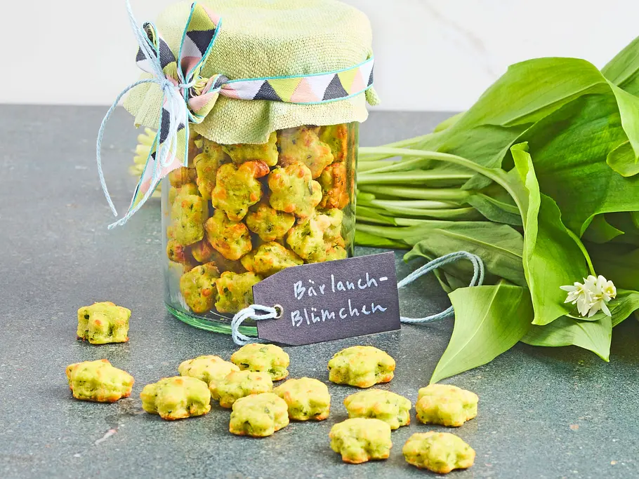 Bärlauch-Blümchen Rezept Kleine, gebackene Bärlauchkekse in Blümchenform, dekorativ in ein Glas mit grünem Stoffdeckel gefüllt, daneben frischer Bärlauch auf einer dunklen Oberfläche.