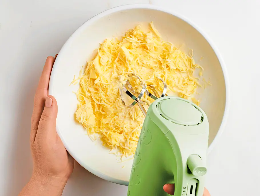 Geriebener Bergkäse, weiche Butter und Sahne werden in einer weißen Schüssel mit einem grünen Handmixer verrührt.