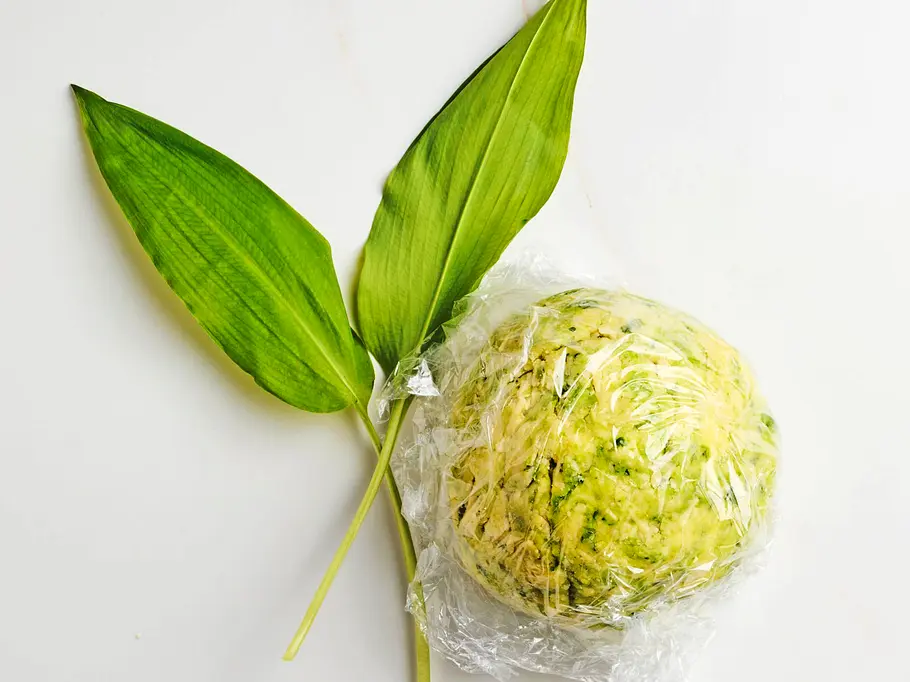Zu einer Kugel geformter Bärlauchteig, in Frischhaltefolie gewickelt und bereit zum Kühlen, daneben frische Bärlauchblätter auf heller Arbeitsfläche.