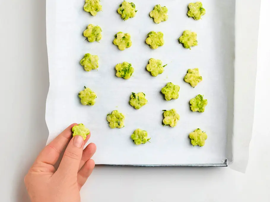 Kleine, grüne Bärlauchblümchen-Kekse auf einem mit Backpapier belegten Blech, bereit zum Backen, eine Hand setzt einen Keks dazu.