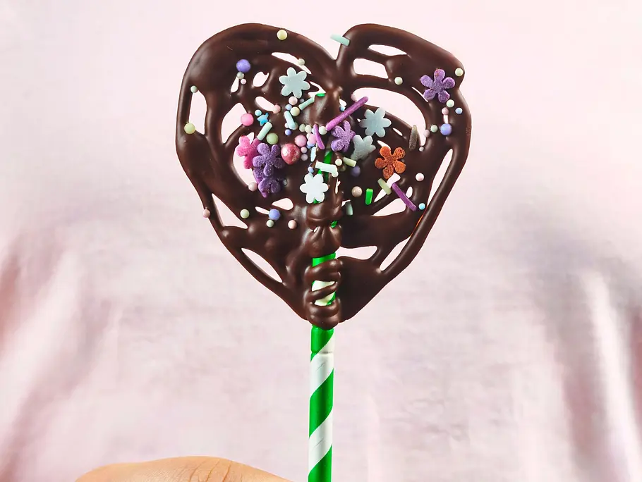 Herzförmiger Schokoladenlolli auf einem grün-weiß gestreiften Stiel, dekoriert mit bunten Zuckerstreuseln in Blütenform.