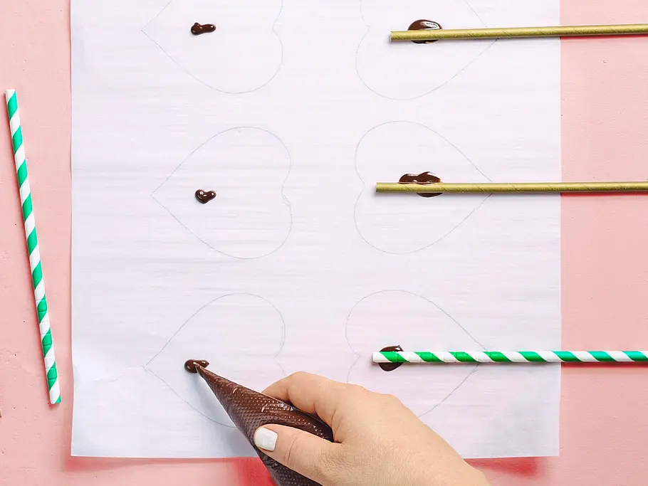 Papier mit vorgezeichneten Herzformen, auf das mit einer Spritztüte Schokolade aufgetragen und Papiertrinkhalme aufgelegt werden.