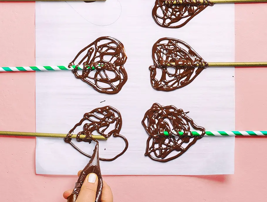 Herzformen aus Schokolade werden mit einer Spritztüte filigran auf Papier gespritzt, Trinkhalme sind als Stiele auf dem Papier platziert.