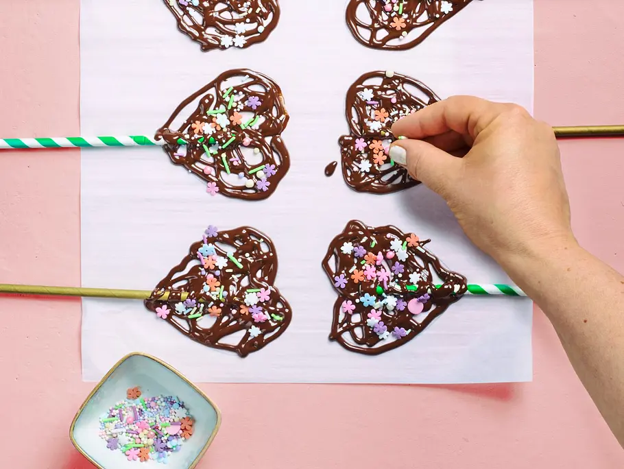 Herzförmige Schokoladenlollis auf Papier, die mit bunten Zuckerstreuseln bestreut werden; daneben eine kleine Schale mit weiterer Streudeko.