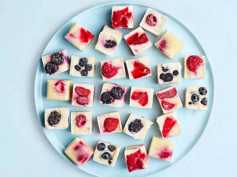 Verschiedene gefrorene Frozen-Yogurt-Würfel mit bunten Beeren auf einem runden, hellblauen Teller.