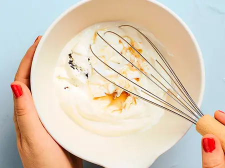 Eine Person verrührt griechischen Joghurt, Honig und Vanille in einer weißen Schüssel mit einem Schneebesen.