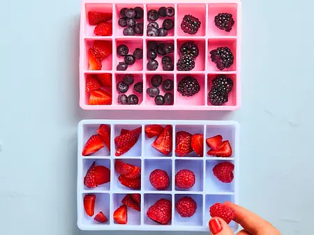 Eine Person verteilt verschiedene Beeren in die Mulden von zwei Silikoneiswürfelformen, eine rosa und eine hellblaue Form.