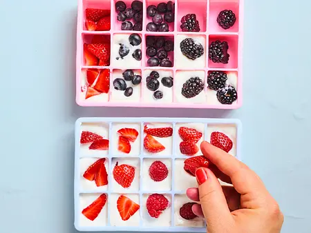 Eine Person drückt Beeren in mit Joghurt gefüllte Mulden zweier Silikoneiswürfelformen, eine rosa und eine hellblaue Form.