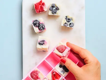 Eine Person löst gefrorene Frozen-Yogurt-Würfel mit Beeren aus einer rosa Eiswürfelform auf einer kleinen weißen Platte.