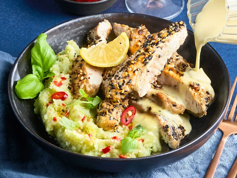 Knuspriges Low-Carb-Tonkatsu auf cremigem Avocado-Blumenkohl-Mash in einer dunklen Schale, garniert mit Zitronenscheibe, Chili und frischem Basilikum; daneben goldenes Besteck auf einem blauen Stoff.