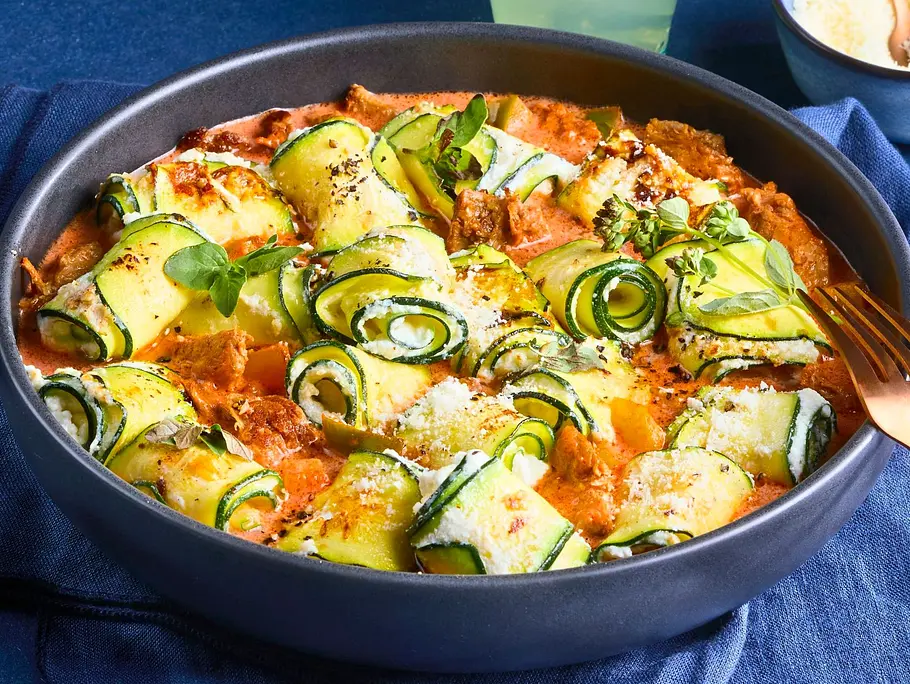 Zucchiniröllchen gefüllt und auf Pulled-Pork-Sugo in einer dunklen Schale angerichtet, serviert mit goldenem Besteck auf dunkelblauer Serviette. Daneben kleine Schale mit Parmesan und ein Glas mit Limetten-Getränk.