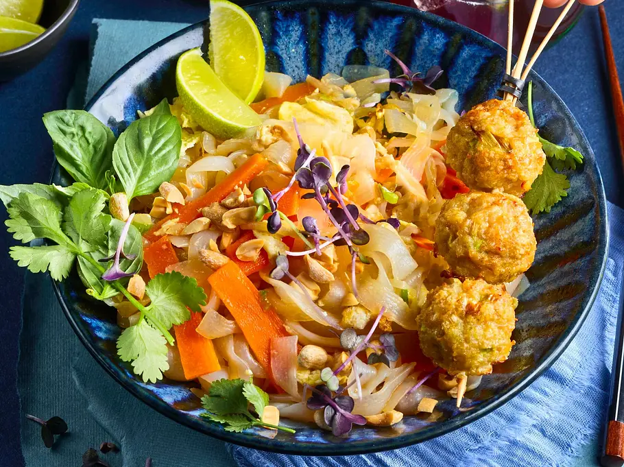 Low Carb-Pad Thai aus Gemüsenudeln, bunten Streifen, Kräutern und gehackten Erdnüssen, serviert mit Geflügelhackbällchen am Spieß auf einem blau gemusterten Teller und platzierter heller Serviette.