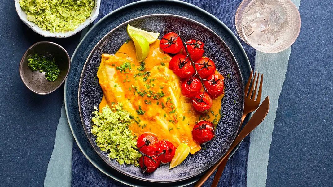 Zart gebratene Asia-Scholle mit gelber Sauce, serviert mit grünem Brokkoli-Reis und gebratenen Kirschtomaten auf dunklem Keramikteller; dazu goldfarbenes Besteck auf dunkelblauer Stoffserviette. - Foto: LECKER @ Bauer Media Group