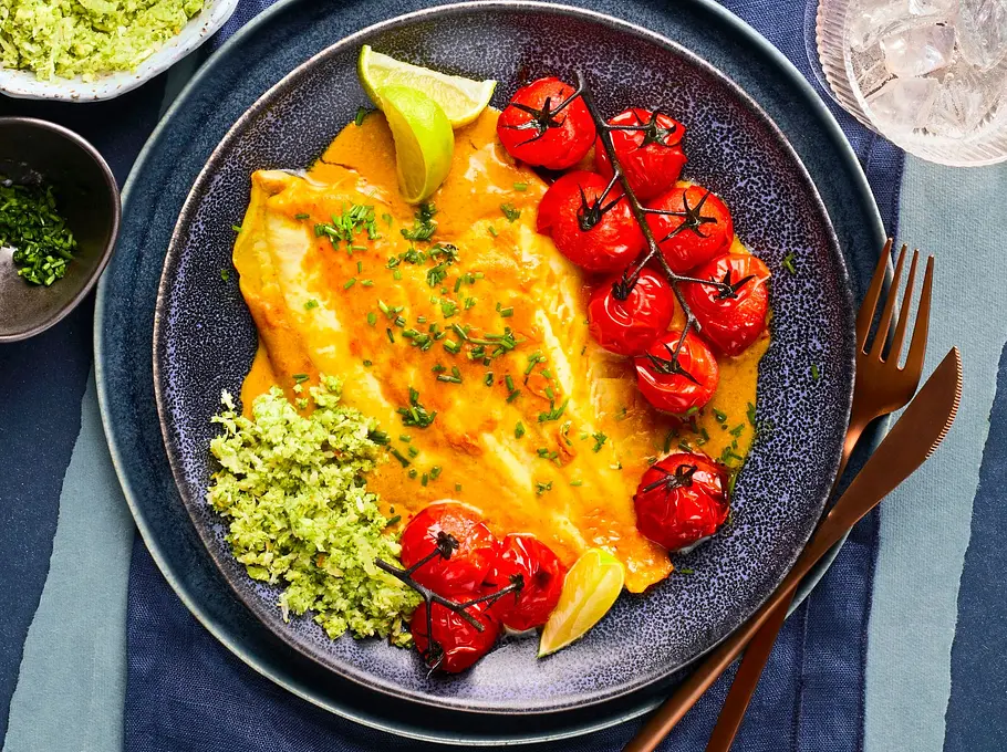 Zart gebratene Asia-Scholle mit gelber Sauce, serviert mit grünem Brokkoli-Reis und gebratenen Kirschtomaten auf dunklem Keramikteller; dazu goldfarbenes Besteck auf dunkelblauer Stoffserviette.