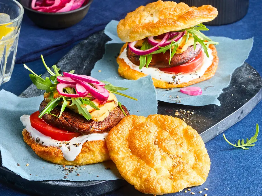 Cloud Bread Burger mit Portobello-Patty Zwei Cloud Bread Burger auf Schieferplatte mit Portobello-Pilz, Tomate, Rucola, roten Zwiebeln und cremigem Dip, serviert auf blauen Papierbögen. Daneben ein Glas mit Zitronenwasser und eine kleine Schale mit eingelegten Zwiebeln.