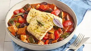 Eine Schale mit buntem, mediterranem Ratatouille-Gemüse und saftig gebratenem Kabeljau-Filet, garniert mit frischen Kräutern. Die Bowl steht auf einer blauen Serviette, daneben liegen Besteck und knuspriges Kräuterbaguette auf einem Holzbrett. - Foto: LECKER @ Bauer Media Group