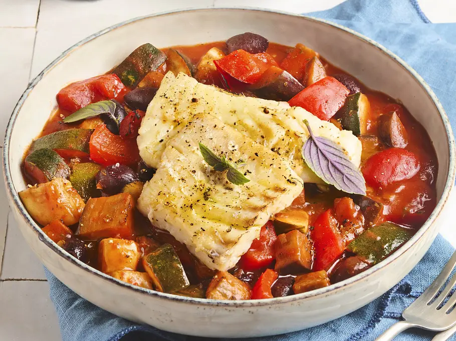 Eine Schale mit buntem, mediterranem Ratatouille-Gemüse und saftig gebratenem Kabeljau-Filet, garniert mit frischen Kräutern. Die Bowl steht auf einer blauen Serviette, daneben liegen Besteck und knuspriges Kräuterbaguette auf einem Holzbrett.