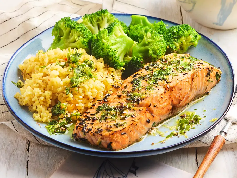 Saftiges Lachsfilet mit Zitronen-Kräuter-Note, dazu zarter Brokkoli und lockerer Bulgur auf einem runden, blauen Teller; das Besteck liegt daneben auf einer gemusterten Fliese.