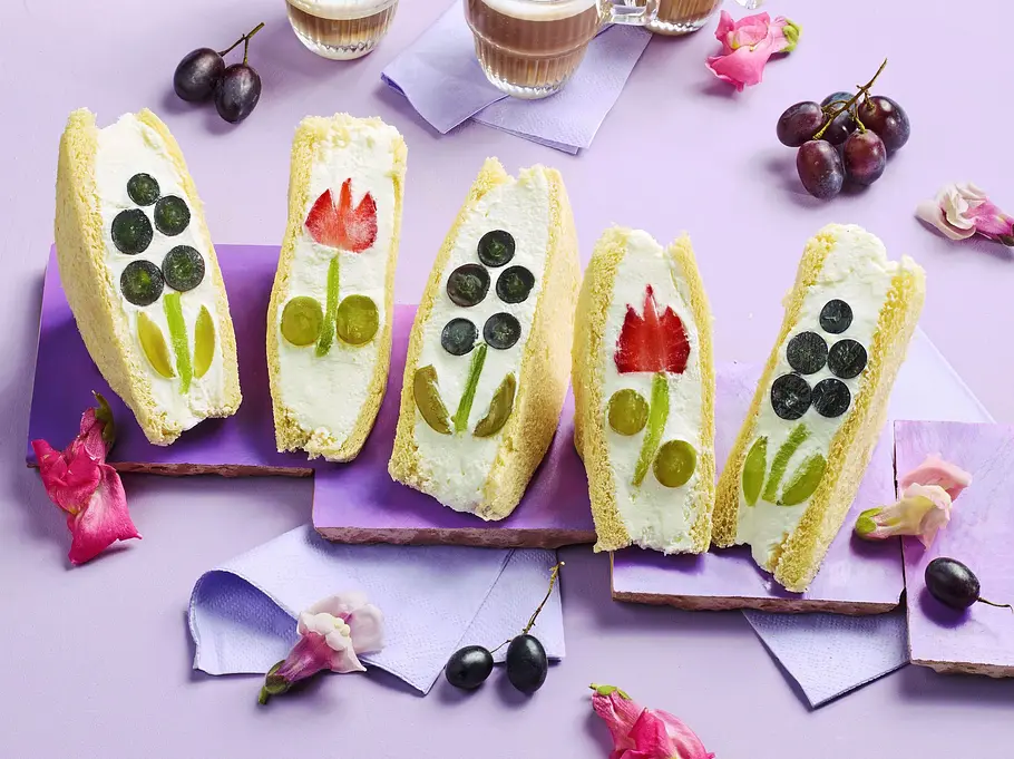 Brioche-Sandwiches, gefüllt mit Creme und frischem Obst wie Trauben und Erdbeeren, kunstvoll zu Blumenmotiven arrangiert, auf pastellfarbenen Unterlagen serviert.