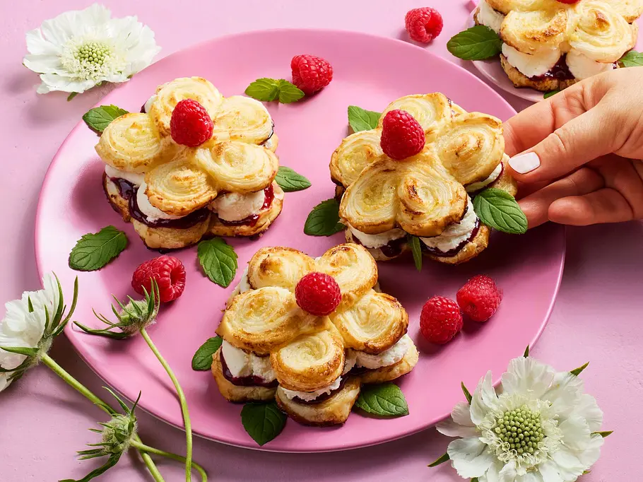 Blätterteig-Gebäcke in Blumenform auf einem rosa Teller, gefüllt mit Sahne und Beeren, dekoriert mit frischen Himbeeren und Minzblättern.