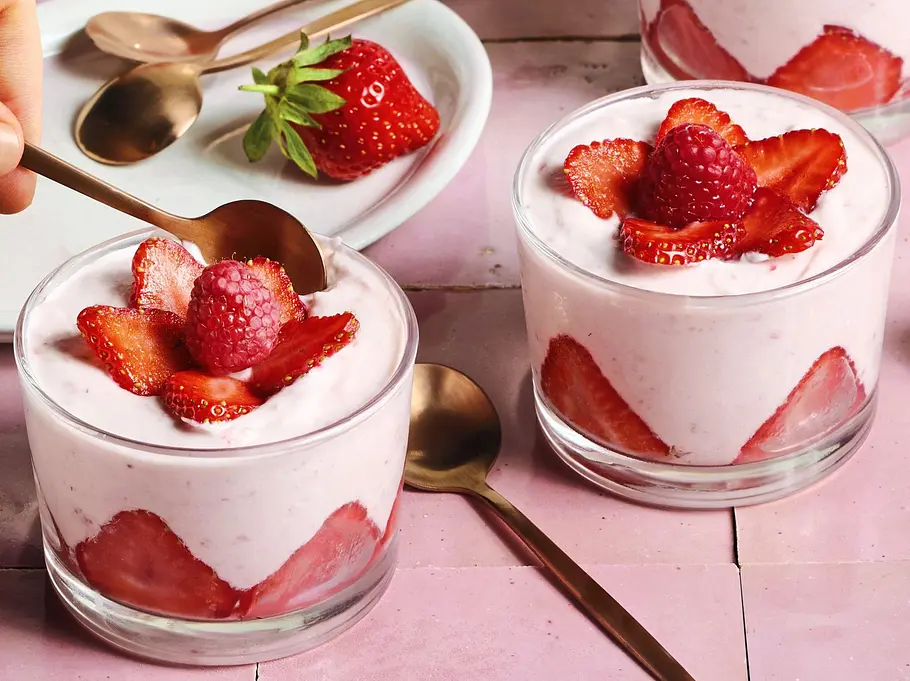 Einfache Erdbeer-Mousse im Glas Vier Gläser mit cremiger Erdbeer-Mousse, dekoriert mit frischen Erdbeerscheiben und einer Himbeere, auf einem rosa Untergrund; daneben liegen weitere Erdbeeren und goldene Löffel.