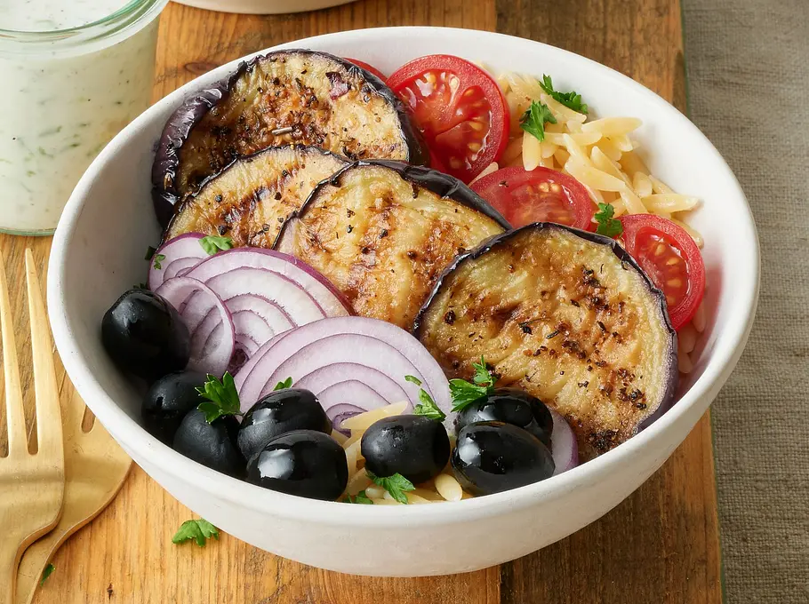 Auberginen-Bowl aus dem Airfryer Rezept Zwei weiße Schalen mit gegrillten Auberginenscheiben, roten Zwiebeln, schwarzen Oliven, Kirschtomaten und Pasta auf einem Holzbrett, daneben ein Glas mit heller Joghurtsoße und goldfarbenes Besteck.