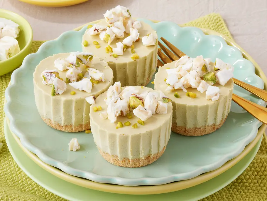 Pistazien-Eistörtchen mit Keksboden Rezept Drei Pistazien-Eistörtchen mit hellem Keksboden, getoppt mit gehackten Pistazien und weißen Stückchen, angerichtet auf einem türkisfarbenen Teller mit goldenen Dessertgabeln auf einem gelbgrünen Platzdeckchen.