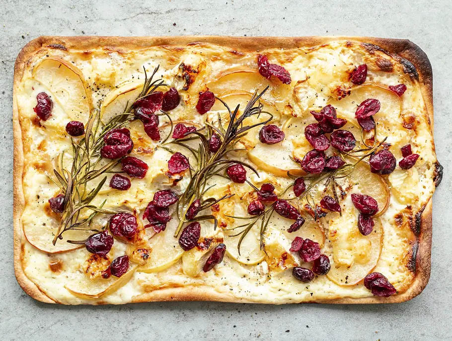 Rechteckiger Flammkuchen auf heller Steinplatte, belegt mit Apfelscheiben, geschmolzenem Brie und getrockneten Cranberrys.
